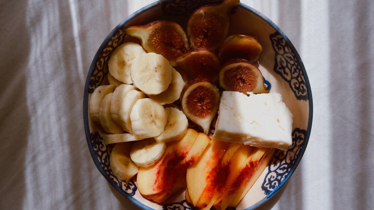 A vibrant bowl of banana, fig, peach, and cheese in decorative dish under sunlight shadows.